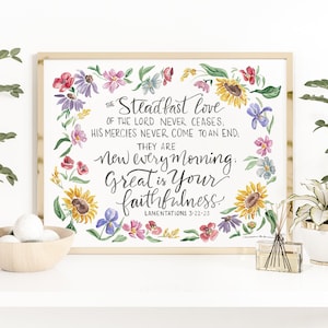 May include: A framed watercolor print with the text "The Steadfast love of the Lord never ceases...Great is Your faithfulness." The artwork is surrounded by a floral wreath of sunflowers, daisies, and other colorful flowers. The frame is a light wood color.