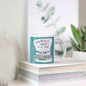 May include: White ceramic mug with a teal background and a black and white illustration of a vintage typewriter. The text "Your story is meant to be told" is written in black cursive on the mug.