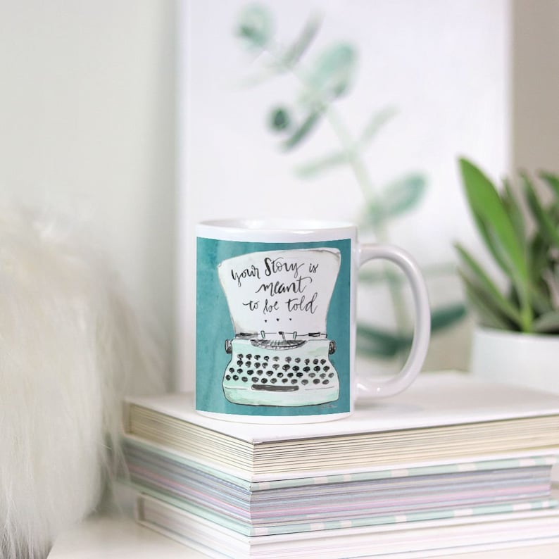 May include: White ceramic mug with a teal background and a black and white illustration of a vintage typewriter. The text "Your story is meant to be told" is written in black cursive on the mug.