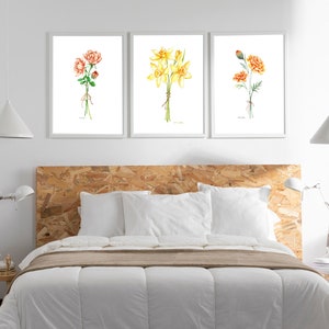 Puede incluir: Tres acuarelas enmarcadas de ramos de flores. La primera pintura presenta un ramo de rosas rosas, la segunda presenta un ramo de narcisos amarillos y la tercera presenta un ramo de claveles naranjas. Las pinturas se exhiben en una pared blanca sobre una cabecera de madera.