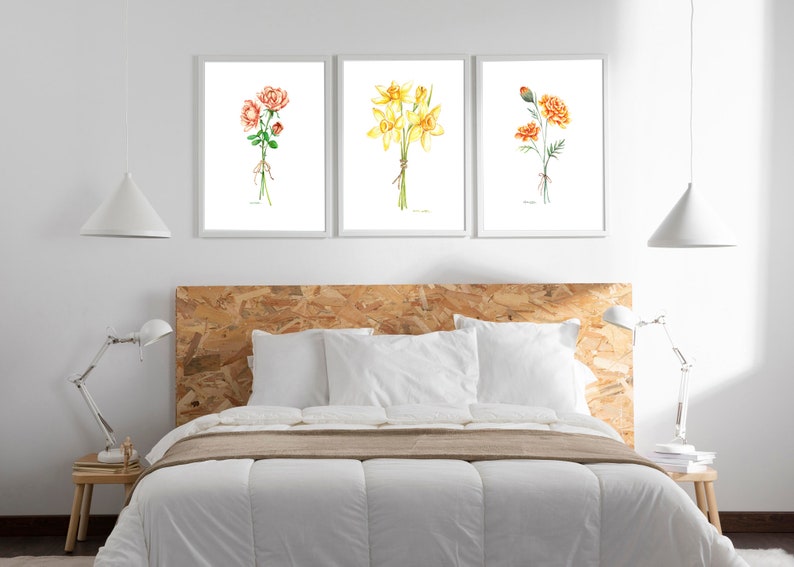May include: Three framed watercolour paintings of bouquets of flowers. The first painting features a bouquet of pink roses, the second features a bouquet of yellow daffodils, and the third features a bouquet of orange carnations. The paintings are displayed on a white wall above a wooden headboard.
