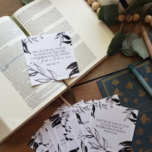 May include: A stack of white cards with black floral designs and inspirational quotes, including "But as for me, I watch in hope for the Lord, I wait for God my Savior; my God will hear me." The cards are on a wooden surface with an open book.