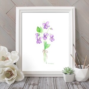 Puede incluir: Una pintura de acuarela de un ramo de violetas moradas con hojas verdes y un lazo de cuerda marr&oacute;n. La pintura est&aacute; enmarcada en un marco blanco y se muestra sobre una superficie de madera.