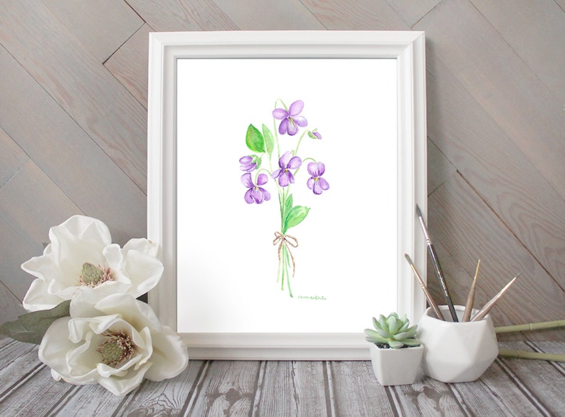 Puede incluir: Una pintura de acuarela de un ramo de violetas moradas con hojas verdes y un lazo de cuerda marr&oacute;n. La pintura est&aacute; enmarcada en un marco blanco y se muestra sobre una superficie de madera.