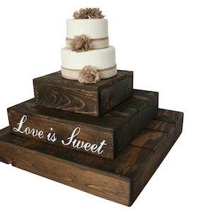 May include: Three-tiered wooden cake stand with a white cake on top. The stand is stained dark brown and has the words "Love is Sweet" written on the bottom tier.