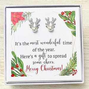 May include: A white gift box with a Christmas theme. The box contains a pair of silver reindeer earrings with pearl accents. The card inside reads "It's the most wonderful time of the year. Merry Christmas!"