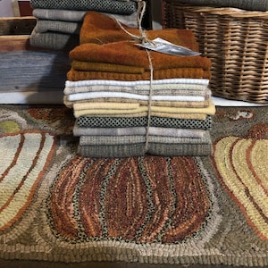 May include: A stack of wool fabric scraps in various shades of brown, beige, and gray, tied together with twine. The fabric is arranged on a rug with a brown and beige pumpkin design.
