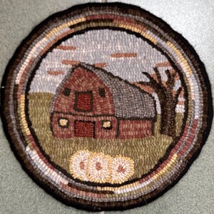 May include: A round, hand-hooked rug featuring a rustic barn scene. The barn is red with a gray roof, set against a cloudy sky and a brown tree. The rug has a circular border with various earth-tone colors, and three pumpkins are at the bottom.