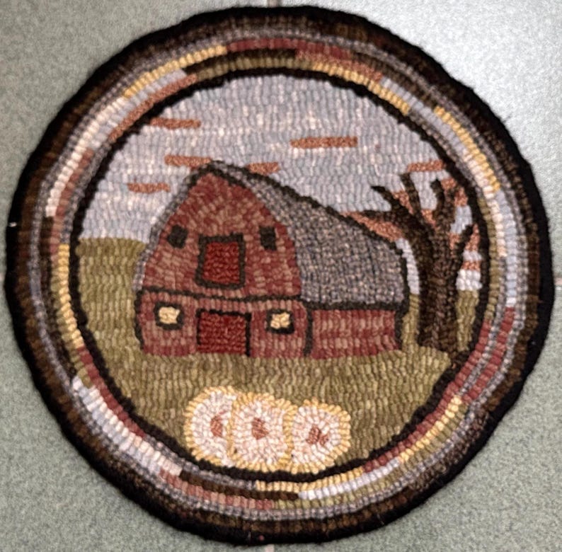 May include: A round, hand-hooked rug featuring a rustic barn scene. The barn is red with a gray roof, set against a cloudy sky and a brown tree. The rug has a circular border with various earth-tone colors, and three pumpkins are at the bottom.