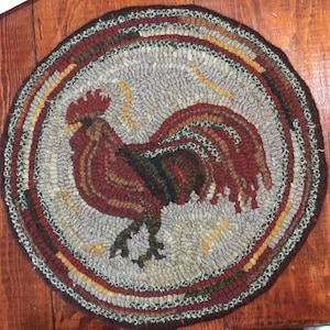 May include: A round wool rug with a rooster design in shades of red, brown, and green. The rooster is surrounded by a border of brown, red, and white wool.