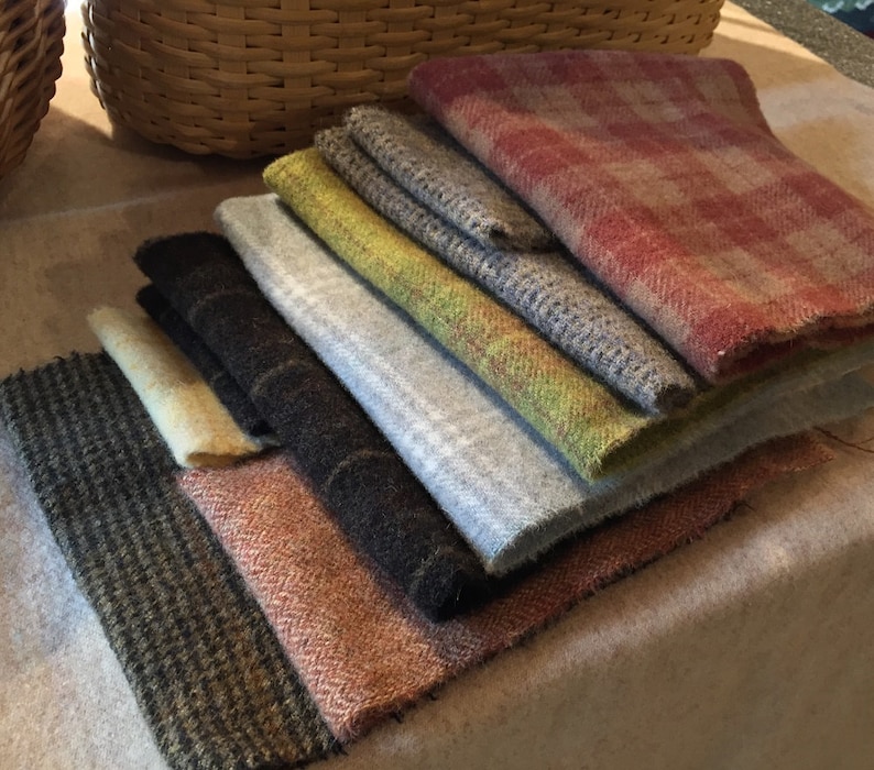 May include: A stack of various wool fabric swatches in shades of gray, brown, green, and red. The fabrics have different textures and patterns, including herringbone and plaid. The swatches are arranged in a fan shape.