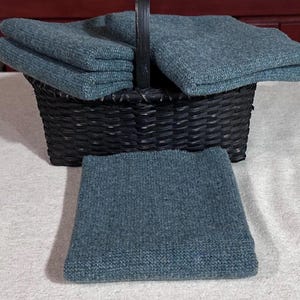 May include: A black wicker basket holds folded teal-colored knitted washcloths. One washcloth rests in front of the basket. The washcloths have a textured appearance. The basket has a handle and is sitting on a white surface.