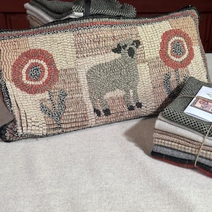 May include: A rectangular hooked rug pillow featuring a central gray sheep standing between two large, circular red and tan flowers. The design is composed of a grid of textured, muted-tone squares. A stack of folded fabric swatches sits nearby.