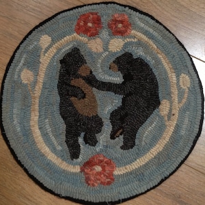 Op de afbeelding: Een rond, blauw en wit tapijt met twee zwarte beren in een cirkel. De beren staan ​​tegenover elkaar en hebben bruine vacht. Er zijn rode bloemen in de hoeken van het tapijt.