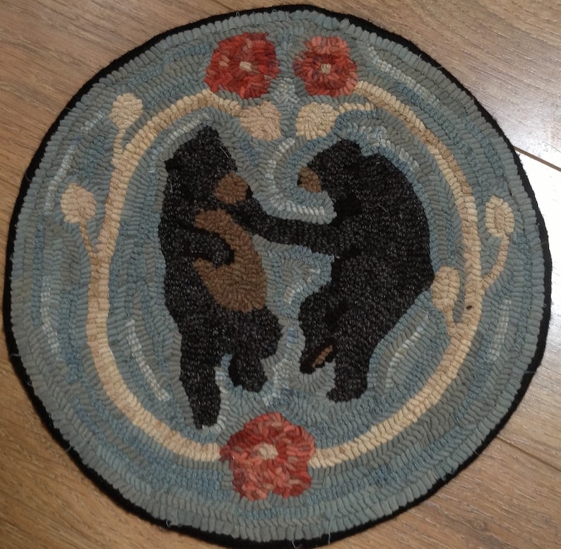 Op de afbeelding: Een rond, blauw en wit tapijt met twee zwarte beren in een cirkel. De beren staan ​​tegenover elkaar en hebben bruine vacht. Er zijn rode bloemen in de hoeken van het tapijt.