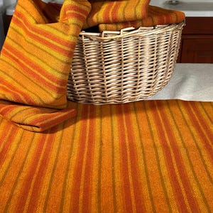 May include: A woven basket holds folded orange and yellow striped textiles. The textiles are arranged on a matching striped fabric. The color palette includes shades of orange, yellow, and brown.