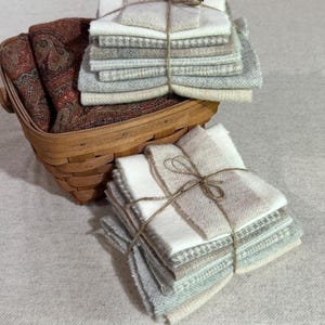 May include: A woven wooden basket containing a stack of folded fabric squares in shades of white, cream, and grey, tied with twine. Beside the basket sits another stack of similar fabric squares, also secured with a simple twine bow.