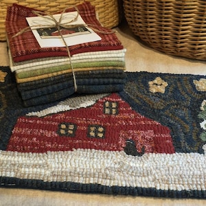 May include: A hand-hooked wool rug depicting a red house with white trim and windows, surrounded by trees and snow. The rug is on a light brown surface with a stack of fabric squares in the background.