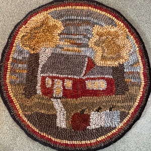 May include: A round, brown, red, and beige hooked rug with a red house, two trees, and a pumpkin in the foreground. The house has three windows and a chimney. The trees are brown and yellow. The pumpkin is orange.