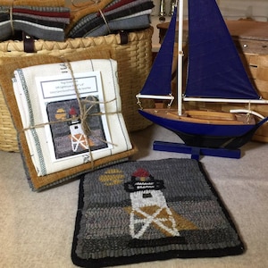 May include: A rug hooking kit featuring a lighthouse design, bundled with fabric strips and a pattern card. A finished square rug with a lighthouse motif sits in front, alongside a model sailboat with a blue hull and dark blue sails.