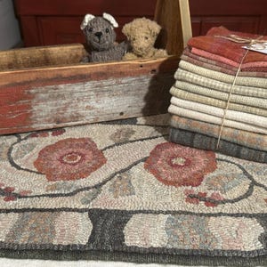 May include: A rectangular rug featuring a hooked floral pattern in shades of pink, gray, and cream, accompanied by a wooden carrier containing two small knitted animal figures and a stack of folded fabric swatches tied with twine.