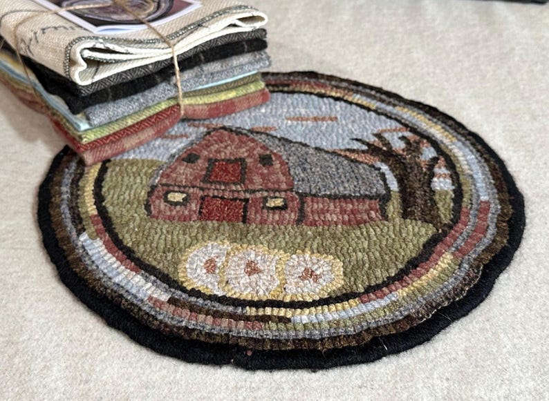 May include: A round, hand-hooked rug featuring a red barn scene with a gray roof, a tree, and a light blue sky. The rug has a black border and a stack of fabric squares tied with twine.