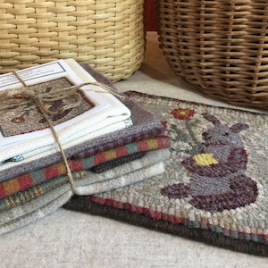 May include: A stack of hand-stitched wool rug hooking supplies, including a finished rug with a bunny design, a pattern, and various fabric pieces. The colors include beige, brown, red, and green. Two woven baskets are in the background.