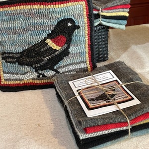 May include: A rug hooking kit featuring a black bird with a red breast. The kit includes wool yarn, a pattern, and instructions. The kit is packaged in a gray fabric bag.