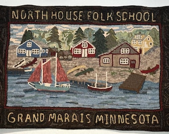 May include: A colorful hooked rug depicting a scene of a small town with houses, trees, and boats. The rug has a brown border with the text "NORTH HOUSE FOLK SCHOOL GRAND MARAIS MINNESOTA" in yellow.