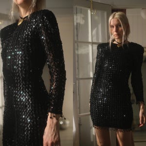 May include: A black sequin dress with long sleeves and shoulder pads. The dress has a round neckline and a fringe detail at the hem. The sequins catch the light, creating a shimmering effect. The dress is knee-length.