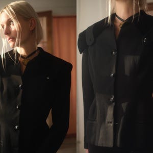 Op de afbeelding: Een zwarte blazer met gestructureerde schouders en een getailleerde taille. De blazer heeft een reverskraag, decoratieve knopen en een getailleerd silhouet. Het kledingstuk is gemaakt van een donkere stof en heeft een vintage uitstraling.