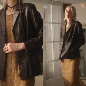 Puede incluir: Una blazer de cuero marrón oscuro con cuello clásico y cierre de botones. La blazer tiene un corte entallado y se lleva sobre un vestido estampado. La blazer tiene un aspecto vintage.