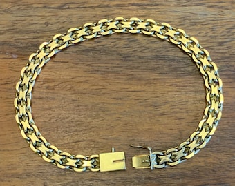 May include: A gold-colored bracelet with a woven chain design. The bracelet is unclasped, revealing the clasp mechanism. The bracelet is lying on a wooden surface.