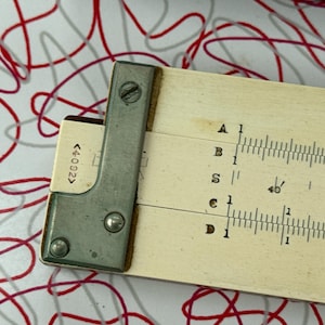 May include: Close-up of a vintage slide rule with a metal frame and a cream-colored scale. The scale has black markings and numbers. The slide rule has the text "<4092>" on the side.