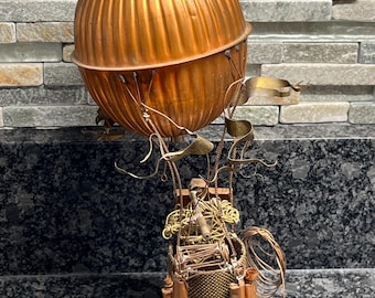 Può includere: Una scultura di mongolfiera in rame con un cesto intrecciato e dettagli intricati in filo metallico. Il pallone è rotondo e ha una texture a coste.