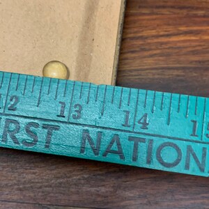 Puede incluir: Primer plano de una regla de color turquesa con las palabras "FIRST NATION" impresas en ella. La regla est&aacute; marcada con n&uacute;meros del 12 al 15. Se puede ver una peque&ntilde;a tachuela redonda de color dorado por encima de la regla.