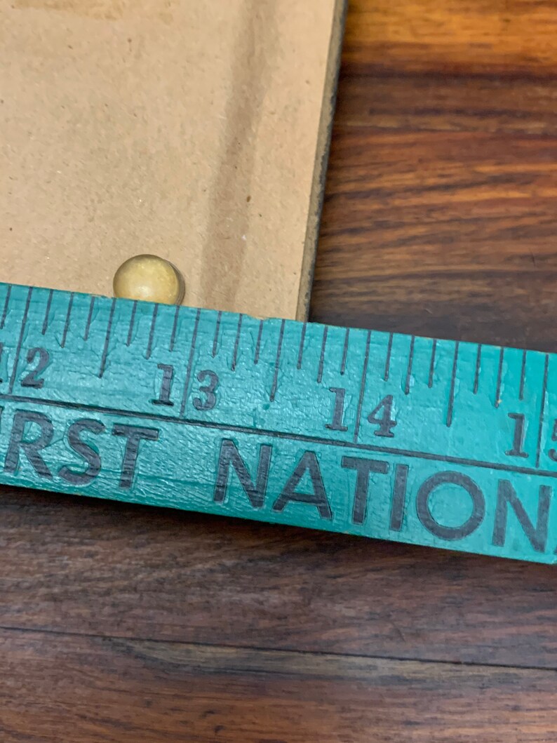 Puede incluir: Primer plano de una regla de color turquesa con las palabras "FIRST NATION" impresas en ella. La regla est&aacute; marcada con n&uacute;meros del 12 al 15. Se puede ver una peque&ntilde;a tachuela redonda de color dorado por encima de la regla.