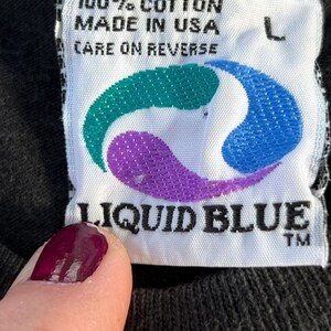 May include: White tag with black text that reads "100% cotton made in USA care on reverse" and a blue, purple, and green logo. Below the logo is the text "LIQUID BLUE TM" in black.