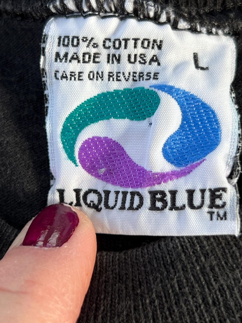 May include: White tag with black text that reads "100% cotton made in USA care on reverse" and a blue, purple, and green logo. Below the logo is the text "LIQUID BLUE TM" in black.