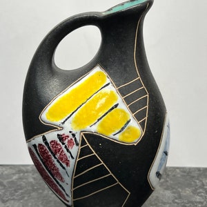 May include: Black ceramic pitcher with a geometric design in yellow, red, white, and blue. The pitcher has a handle and a rounded body.