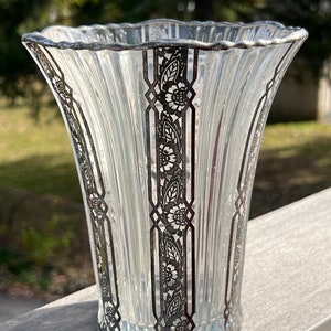 May include: A clear glass vase with a silver floral design. The vase has a scalloped rim and a fluted body.