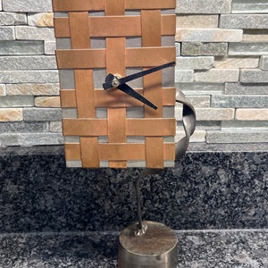 May include: A unique clock with a copper and gray woven face, black hands, and a metal stand. The clock is mounted on a metal base.
