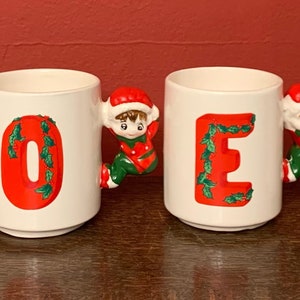 May include: Set of four white ceramic mugs with red letters spelling "NOEL" and green holly leaf accents. Each mug features a small elf figurine holding onto the handle.