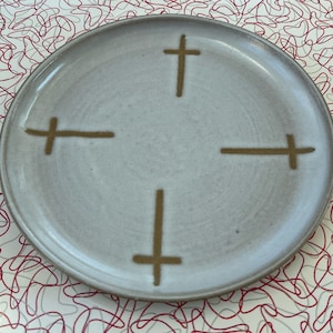 May include: A white ceramic plate with a simple design of four brown crosses. The plate has a slightly textured surface.