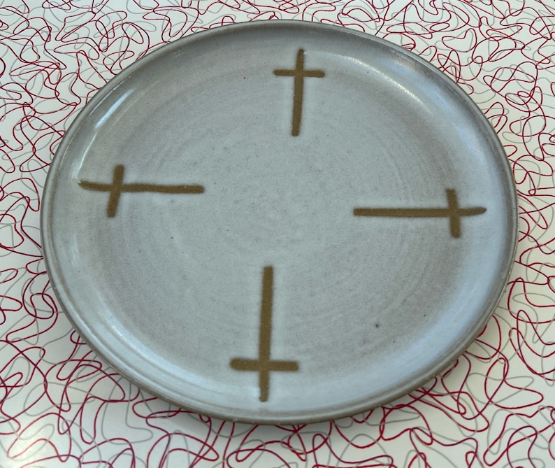 May include: A white ceramic plate with a simple design of four brown crosses. The plate has a slightly textured surface.