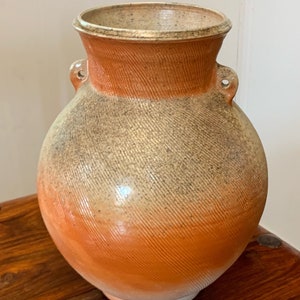 May include: A ceramic vase with a rounded body and a wide mouth. The vase has a light brown glaze with a speckled finish and two small handles on the sides.