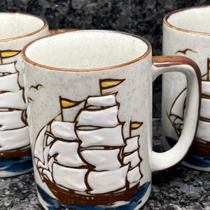 May include: Set of three white ceramic mugs with a brown rim and handle. Each mug features a brown and white illustration of a sailing ship with yellow sails against a blue and white ocean background.