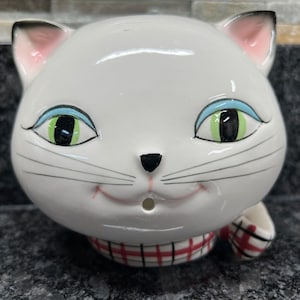 May include: White ceramic cat figurine with blue and green eyes, a pink nose, and a red and white plaid scarf. The cat has a smiling expression and a small hole in its mouth.