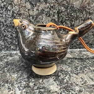 May include: A dark brown glazed ceramic bird-shaped whistle with a glossy finish, featuring incised wing details, a light tan beak, and a circular base. A tan leather cord is threaded through the top of the whistle for hanging or carrying.