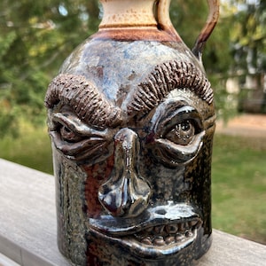 May include: A ceramic jug with a face sculpted on the front. The face has a grumpy expression with furrowed brows, a large nose, and a wide mouth with teeth. The jug is glazed in shades of brown, black, and white.